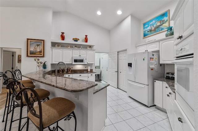 kitchen featuring high vaulted ceiling, white cabinets, white appliances, kitchen peninsula, and a kitchen bar