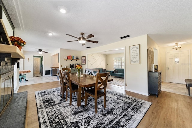 dining room featuring a brick fireplace, a textured ceiling, light wood-type flooring, and ceiling fan