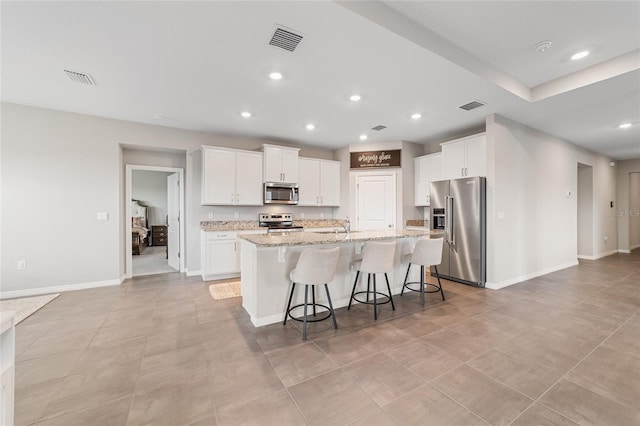 kitchen with white cabinets, stainless steel appliances, light stone counters, and a center island with sink