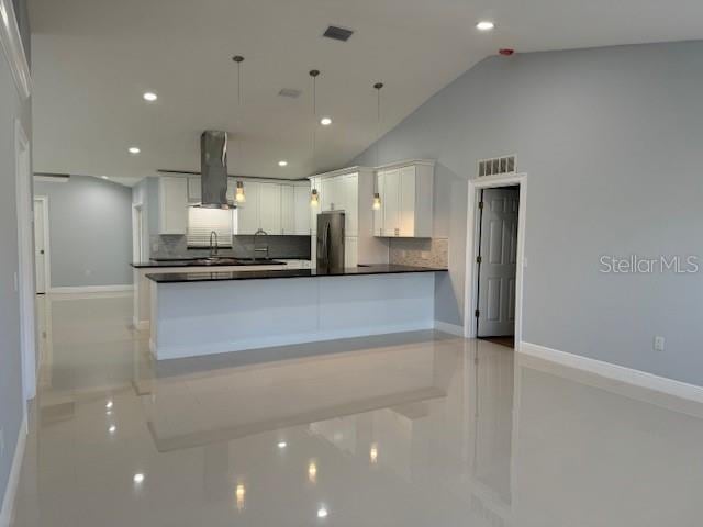 kitchen with wall chimney range hood, lofted ceiling, stainless steel fridge, hanging light fixtures, and white cabinetry