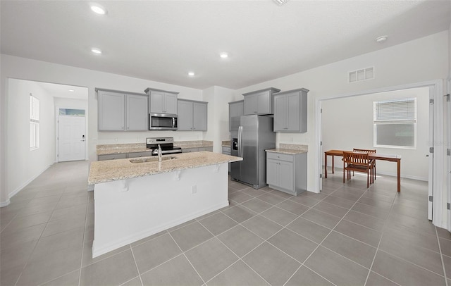 kitchen featuring a center island with sink, a kitchen bar, appliances with stainless steel finishes, gray cabinetry, and light stone counters