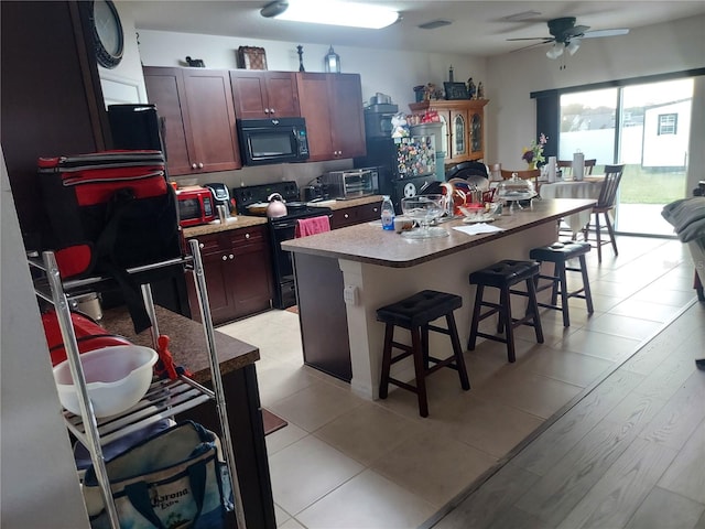 kitchen featuring light wood-type flooring, an island with sink, a kitchen bar, black appliances, and ceiling fan