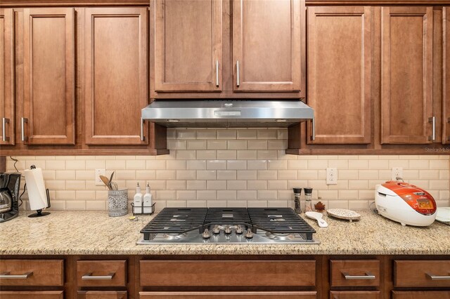 kitchen featuring decorative backsplash, light stone countertops, wall chimney range hood, and stainless steel gas cooktop