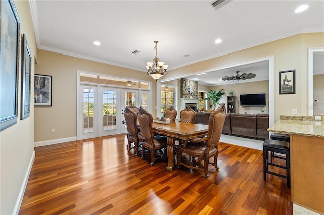 dining space featuring dark wood-type flooring, a stone fireplace, crown molding, and ceiling fan with notable chandelier