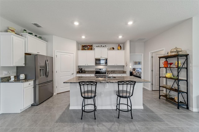 kitchen featuring a textured ceiling, a kitchen island with sink, white cabinets, stainless steel appliances, and dark stone countertops