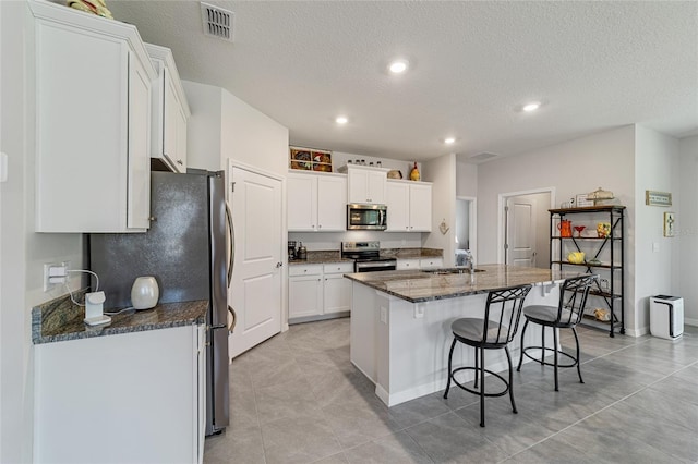 kitchen with dark stone countertops, white cabinetry, stainless steel appliances, a center island with sink, and sink