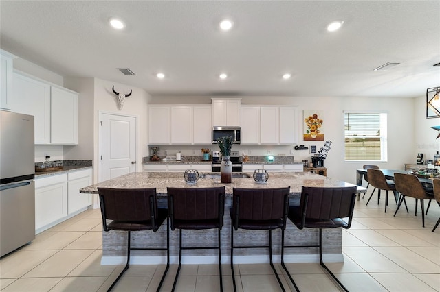 kitchen featuring light stone counters, a kitchen island with sink, white cabinetry, stainless steel appliances, and a kitchen bar