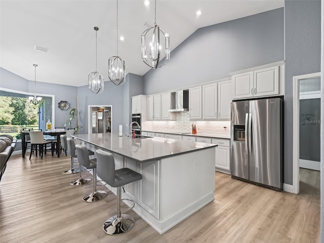 kitchen with white cabinets, wall chimney exhaust hood, stainless steel fridge, and a kitchen island with sink