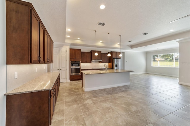 kitchen featuring a center island with sink, stainless steel appliances, light stone countertops, backsplash, and a textured ceiling