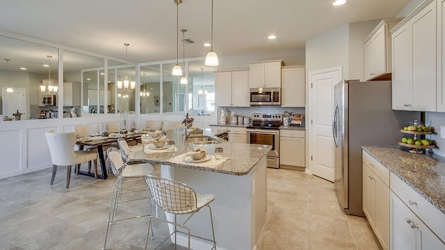 kitchen with light stone counters, stainless steel appliances, white cabinetry, a center island with sink, and decorative light fixtures