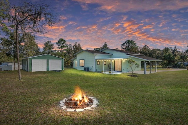 back house at dusk with central AC, a fire pit, a garage, a lawn, and an outdoor structure