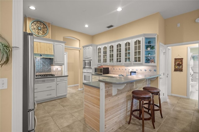 kitchen with light tile patterned floors, a peninsula, glass insert cabinets, appliances with stainless steel finishes, and a kitchen breakfast bar