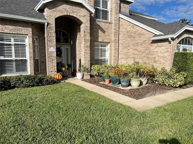 doorway to property featuring a shingled roof, a lawn, and brick siding