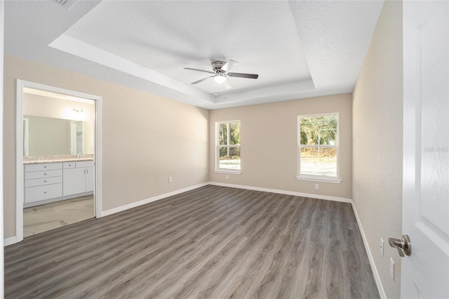 unfurnished bedroom featuring wood-type flooring, a tray ceiling, a textured ceiling, ensuite bathroom, and ceiling fan