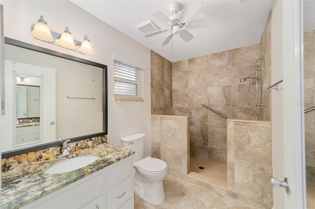 bathroom featuring ceiling fan, a textured ceiling, toilet, tiled shower, and vanity