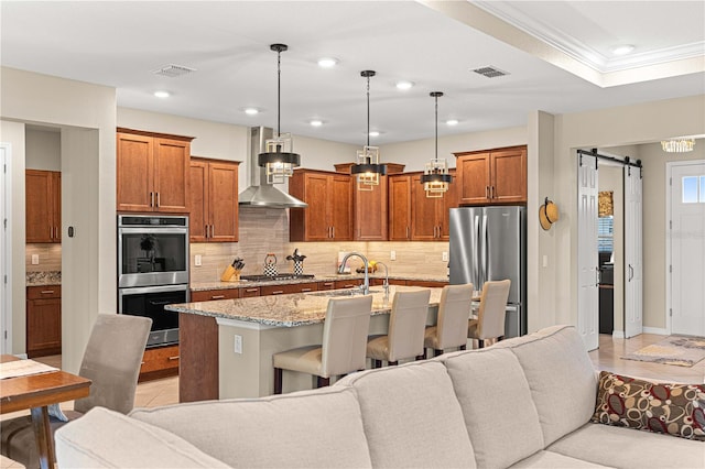 kitchen with a kitchen island with sink, wall chimney range hood, hanging light fixtures, a barn door, and appliances with stainless steel finishes