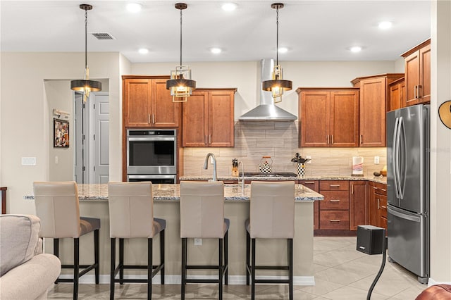 kitchen featuring wall chimney range hood, an island with sink, a kitchen breakfast bar, stainless steel appliances, and decorative light fixtures