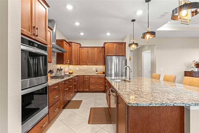 kitchen with a kitchen island with sink, wall chimney exhaust hood, stainless steel appliances, sink, and a kitchen bar