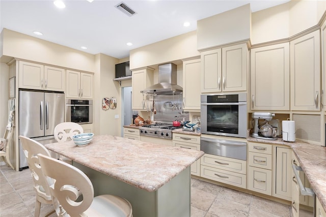 kitchen featuring a breakfast bar area, appliances with stainless steel finishes, cream cabinetry, wall chimney exhaust hood, and light stone counters