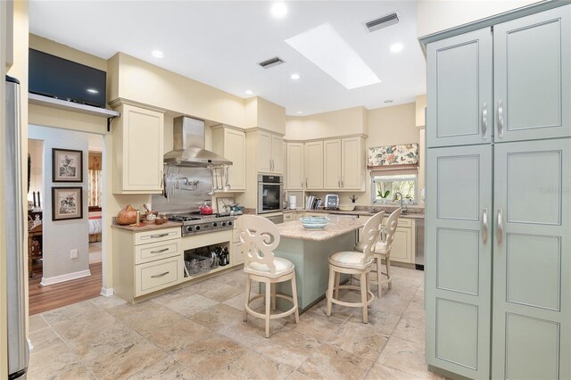 kitchen featuring wall chimney range hood, a skylight, stainless steel appliances, a kitchen island, and a breakfast bar area