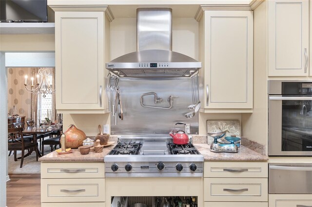 kitchen with wall chimney range hood, hardwood / wood-style floors, appliances with stainless steel finishes, light stone countertops, and a notable chandelier