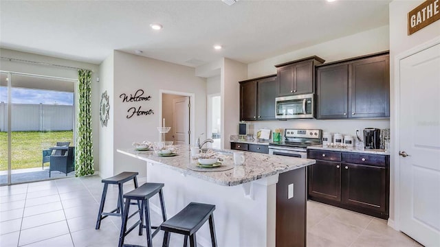 kitchen featuring a breakfast bar, an island with sink, light stone counters, appliances with stainless steel finishes, and light tile patterned floors