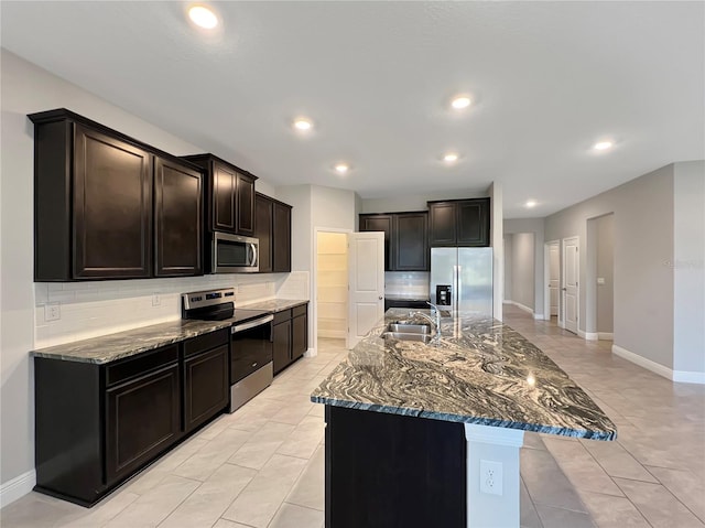 kitchen with backsplash, dark stone countertops, a center island with sink, sink, and appliances with stainless steel finishes