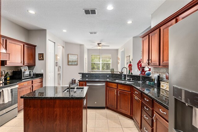 kitchen featuring sink, a textured ceiling, stainless steel appliances, dark stone counters, and a center island with sink