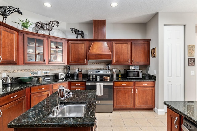 kitchen featuring appliances with stainless steel finishes, sink, dark stone countertops, premium range hood, and a center island with sink