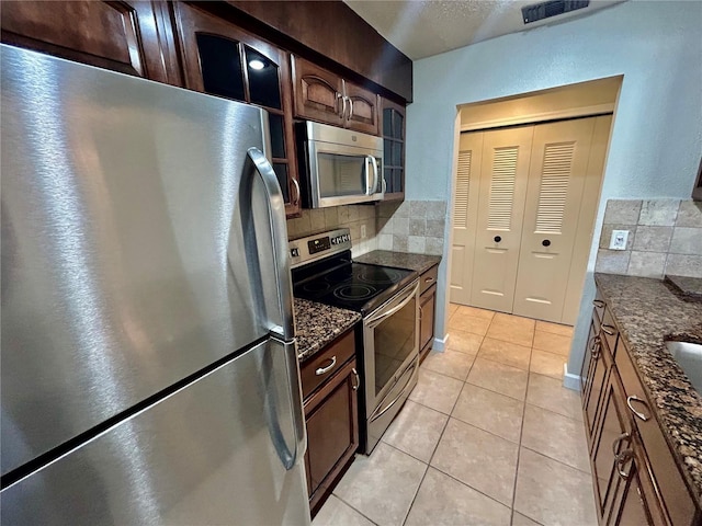 kitchen featuring light tile patterned flooring, backsplash, appliances with stainless steel finishes, dark brown cabinetry, and dark stone countertops