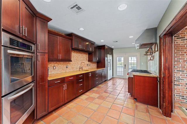 kitchen featuring exhaust hood, light tile patterned floors, appliances with stainless steel finishes, backsplash, and french doors