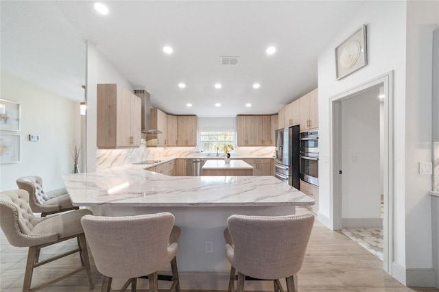 kitchen featuring light brown cabinets, kitchen peninsula, a kitchen breakfast bar, wall chimney exhaust hood, and light stone counters