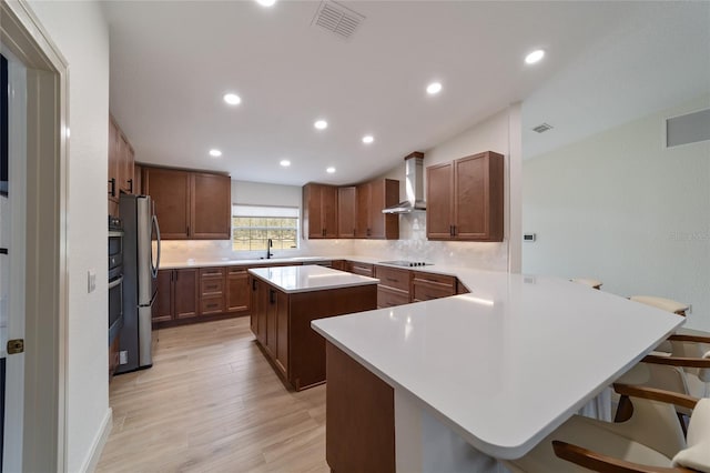 kitchen with a kitchen island, a breakfast bar area, wall chimney exhaust hood, light hardwood / wood-style flooring, and stainless steel refrigerator