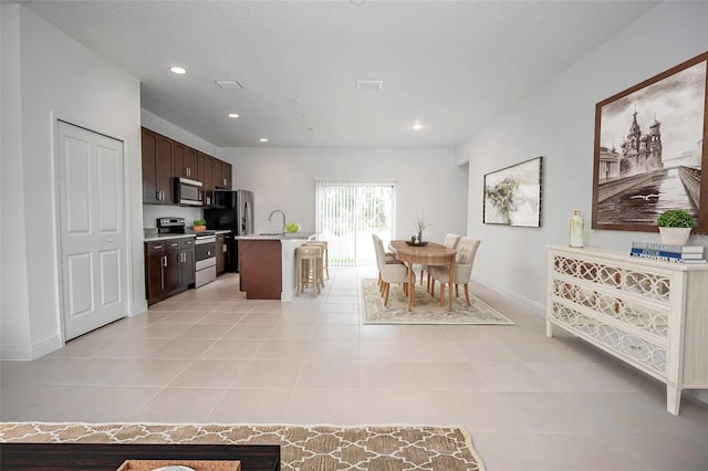 kitchen with an island with sink, dark brown cabinets, sink, light tile patterned flooring, and appliances with stainless steel finishes