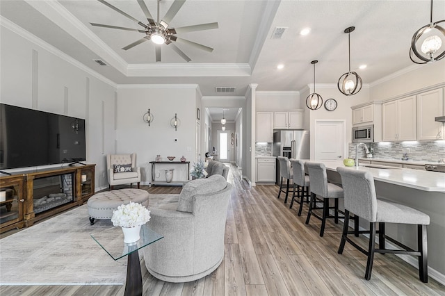 living room with ornamental molding, sink, a tray ceiling, ceiling fan, and light hardwood / wood-style flooring