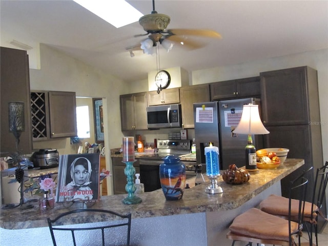 kitchen with kitchen peninsula, dark brown cabinets, vaulted ceiling with skylight, appliances with stainless steel finishes, and ceiling fan