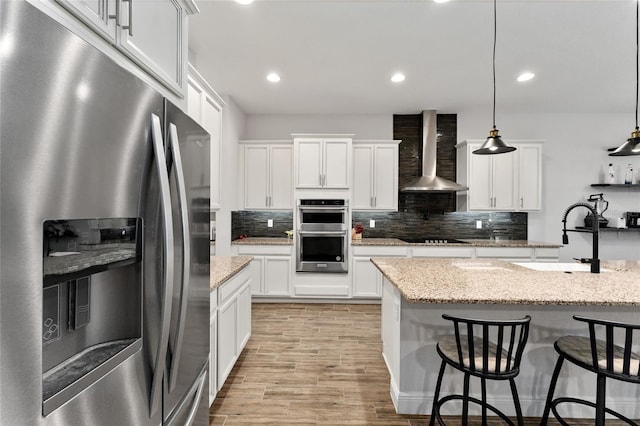 kitchen with sink, appliances with stainless steel finishes, decorative light fixtures, white cabinets, and wall chimney range hood