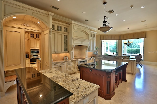 kitchen with appliances with stainless steel finishes, dark stone counters, decorative light fixtures, a kitchen island with sink, and a notable chandelier