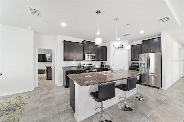 kitchen with a kitchen bar, stainless steel appliances, dark stone counters, a kitchen island with sink, and pendant lighting