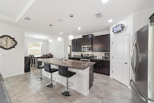 kitchen with stainless steel appliances, dark stone counters, a breakfast bar area, an island with sink, and decorative light fixtures