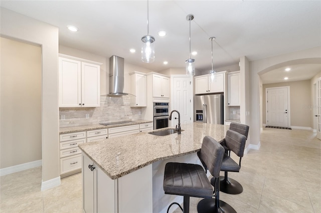 kitchen featuring a center island with sink, white cabinets, wall chimney range hood, light stone countertops, and appliances with stainless steel finishes