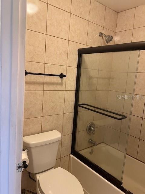 bathroom featuring bath / shower combo with glass door, toilet, and tile walls