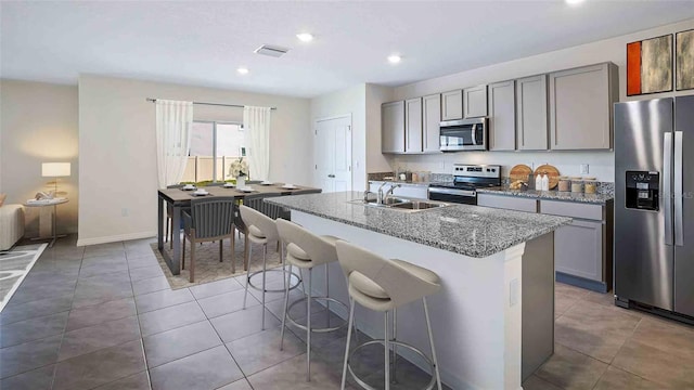 kitchen featuring stainless steel appliances, light stone counters, sink, an island with sink, and gray cabinets