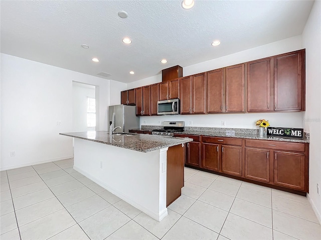 kitchen with sink, dark stone counters, a center island with sink, light tile patterned floors, and appliances with stainless steel finishes