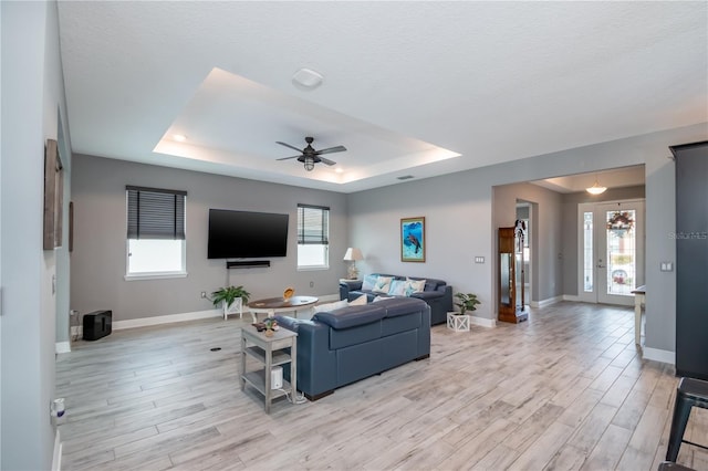 living room with ceiling fan, a textured ceiling, a tray ceiling, light wood-type flooring, and french doors