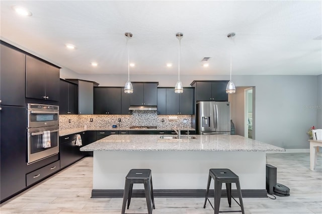 kitchen with light hardwood / wood-style flooring, hanging light fixtures, sink, a kitchen island with sink, and appliances with stainless steel finishes