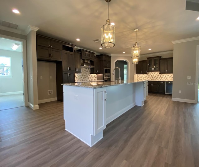 kitchen with arched walkways, dark brown cabinetry, ornamental molding, an island with sink, and decorative light fixtures