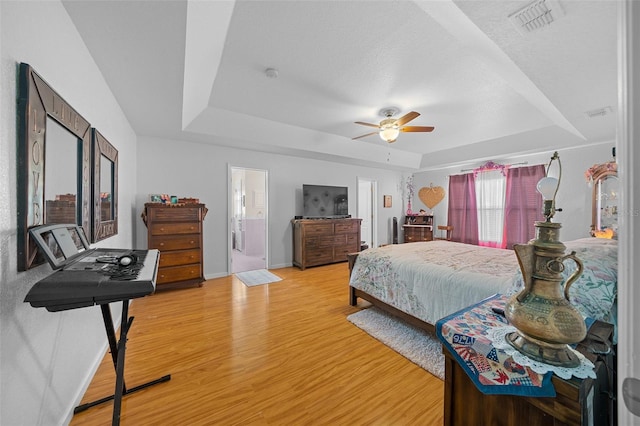 bedroom with ceiling fan, ensuite bathroom, a tray ceiling, and light hardwood / wood-style flooring