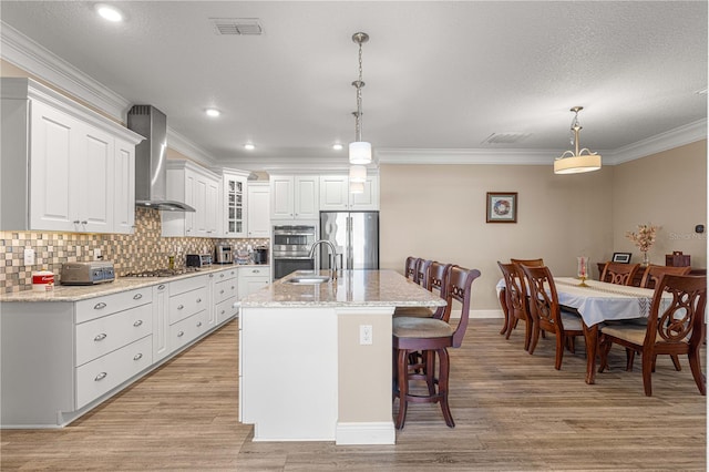 kitchen featuring hanging light fixtures, wall chimney range hood, a kitchen island with sink, and appliances with stainless steel finishes