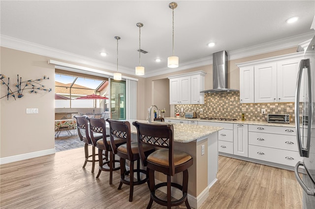 kitchen with white cabinetry, wall chimney exhaust hood, a kitchen island with sink, and decorative light fixtures